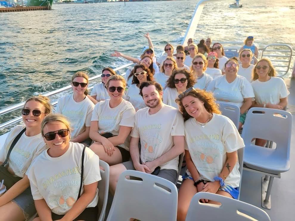 A group of people smiling on a boat with water and a cityscape in the background.