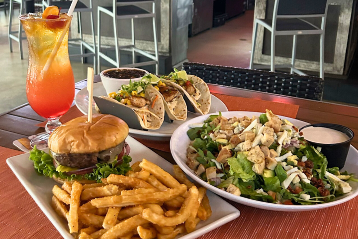 Burger with fries, salad, tacos, and cocktail on a table in a restaurant setting.