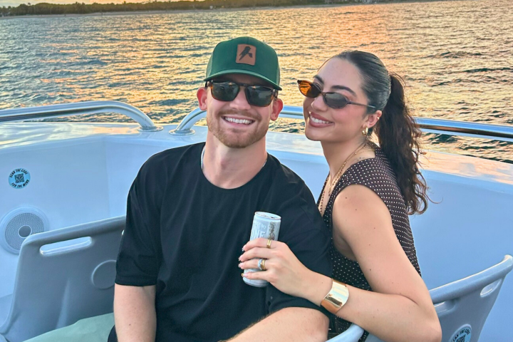 Two people smiling on a boat during sunset, holding a drink can.