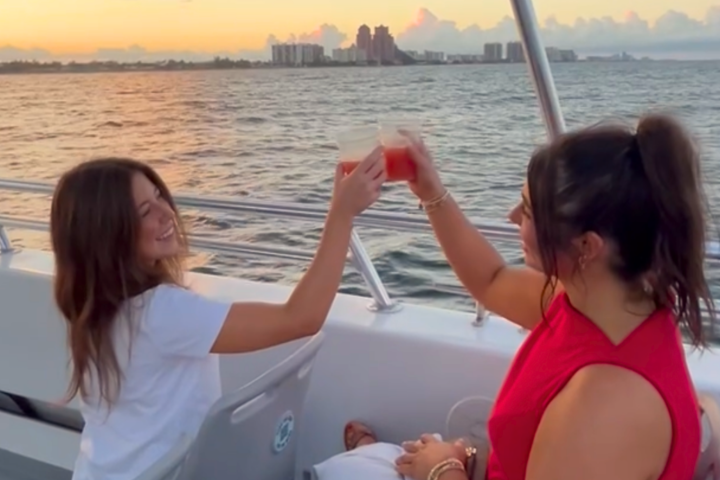 Two women toasting drinks on a boat at sunset with city skyline in the background.