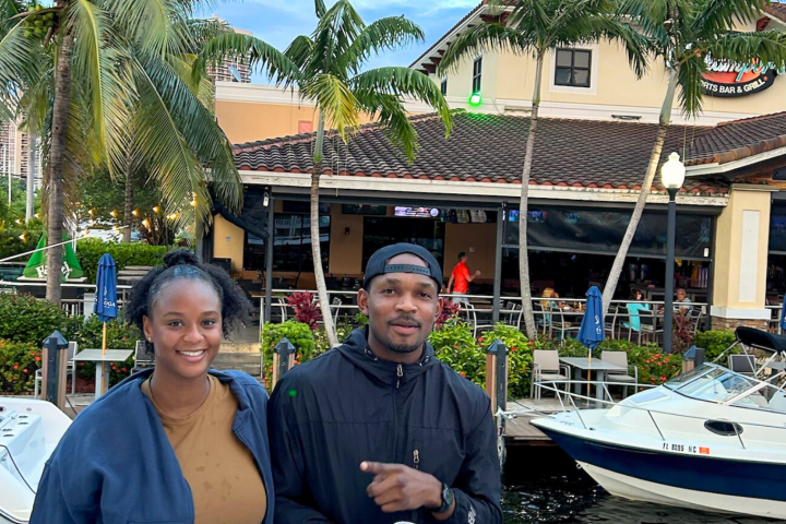 Two people smiling by a docked boat and a tropical-themed restaurant with palm trees.