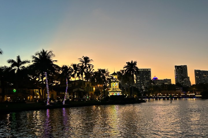 A waterfront cityscape at dusk with palm trees and illuminated buildings reflecting on the water.