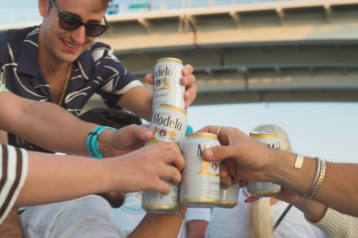Group of people toasting with Modelo beer cans under a bridge.