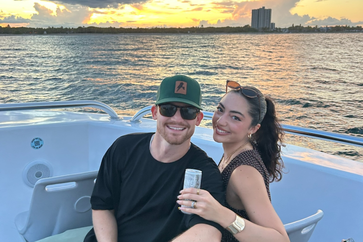 Smiling couple on a boat, with a scenic sunset and city skyline in the background.