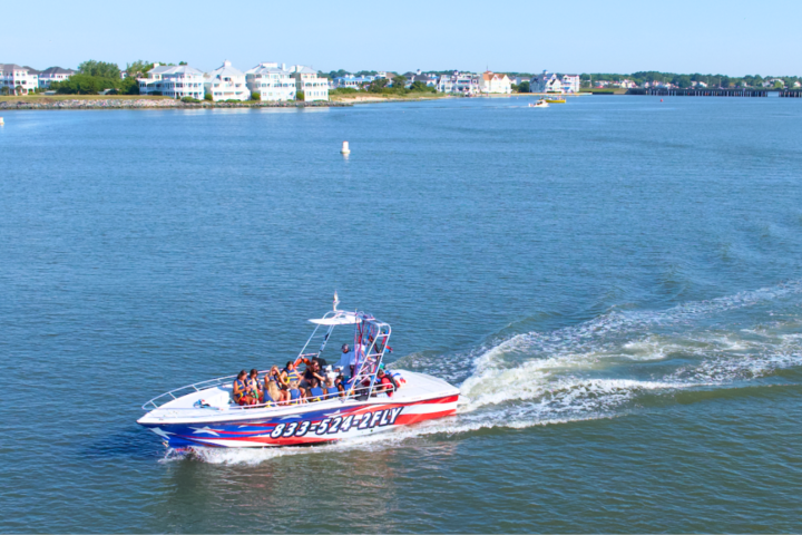 Speedboat with passengers on a calm river, houses and trees in the background.