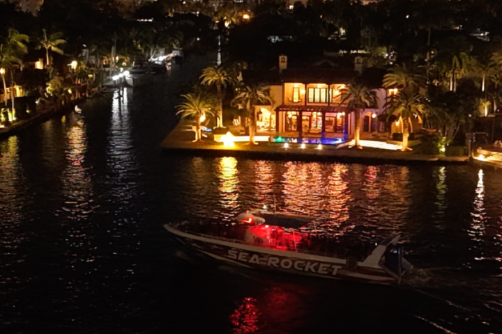 Boat with red lights on canal near large illuminated house at night.