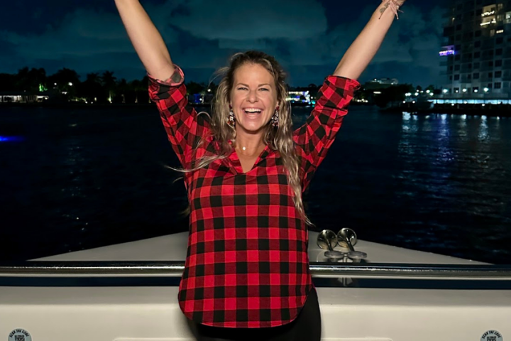 Person in plaid shirt joyfully raising arms on a boat at night with a cityscape in the background.