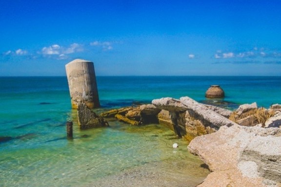 Ruined structures in clear shallow water against a blue ocean and sky.