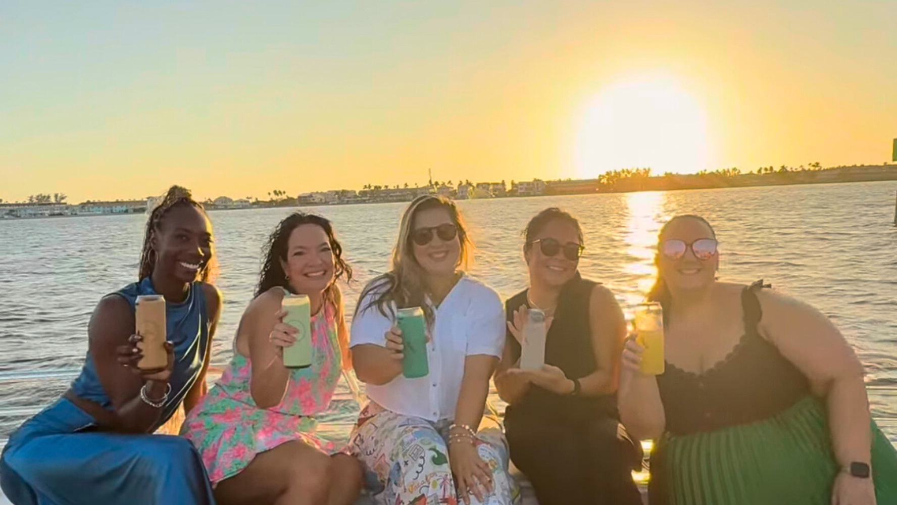 Group of friends holding drinks and smiling on a Bradenton Beach private sunset cruise with golden sunlight over the water.