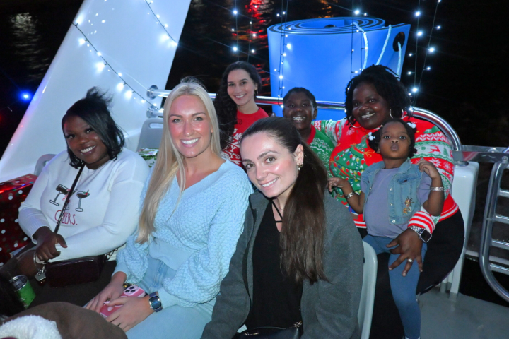 Group of people smiling on a decorated boat with holiday lights and a large gift box.