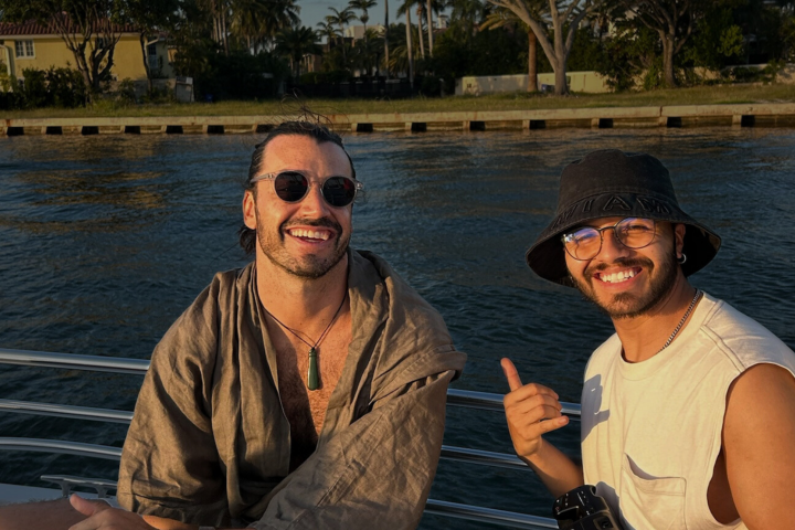Two men smiling on a boat with water and shoreline in the background.