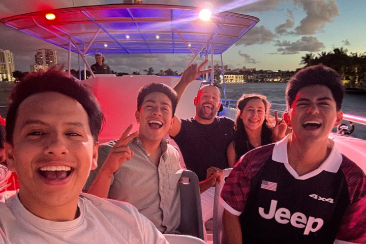 Group of smiling people on a boat at sunset with cityscape in the background.