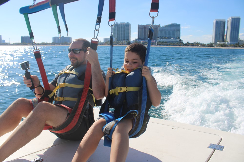 Two people in life jackets parasailing over water with city skyline in background.