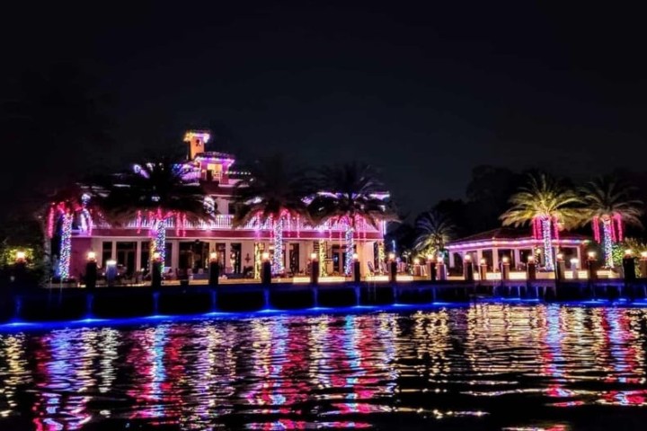 Colorful lights illuminate a house and palm trees, reflecting on water at night.