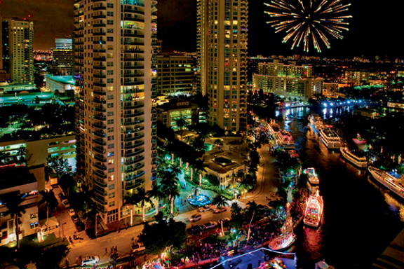Cityscape at night with fireworks, tall buildings, and a parade of illuminated boats on a river.