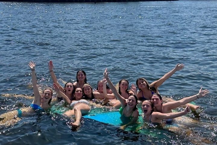 Group of people smiling and waving while floating in water with luxury houses and a yacht in the background.