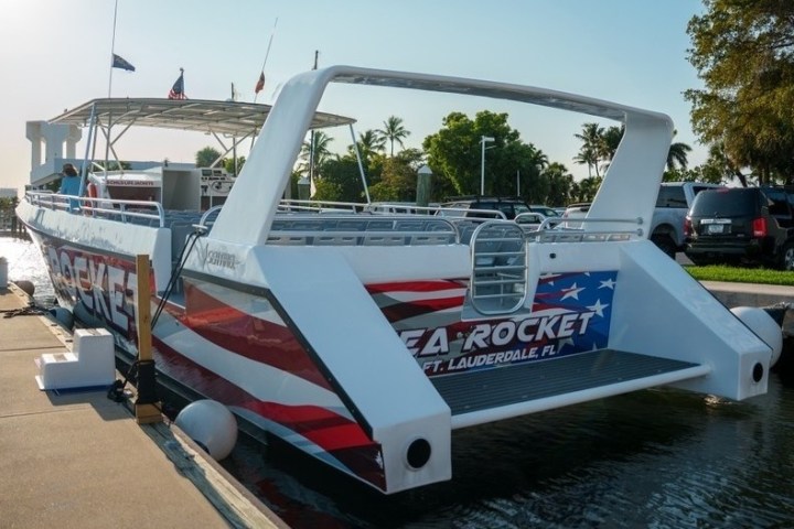 Catamaran docked at marina with 'Sea Rocket' text on stern, palm trees and parked cars in background.