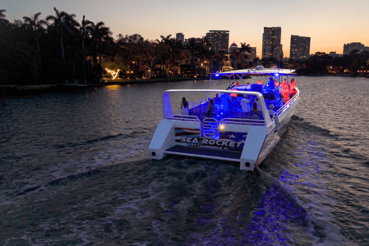 Boat with colored lights on water at sunset with city skyline in background.