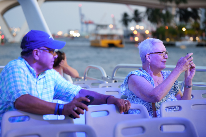 Two people on a boat taking photos, with a cityscape and water in the background.