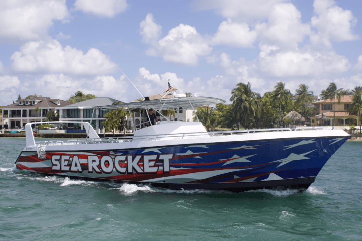 A speedboat named 'Sea Rocket' with a patriotic design cruises near a shoreline with houses and palm trees.