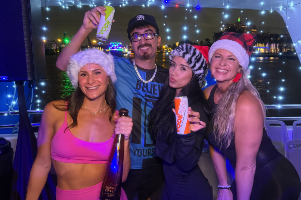 Four people in festive hats smiling and holding drinks on a boat with night lights.