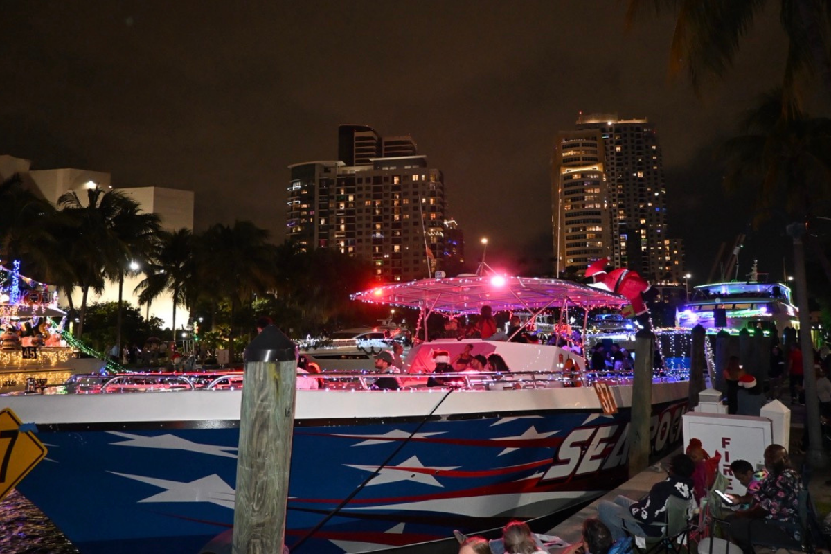 Boat with festive lights and a Santa figure at night, skyscrapers and palm trees in the background.