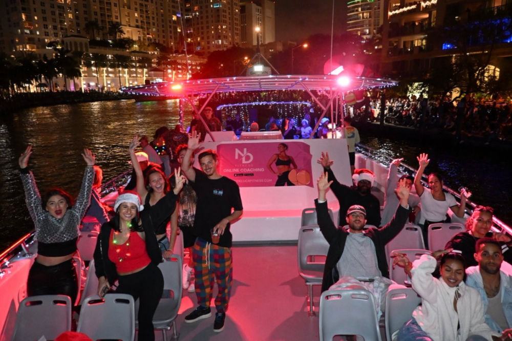 People on a festive boat at night, smiling and waving, with a city backdrop.