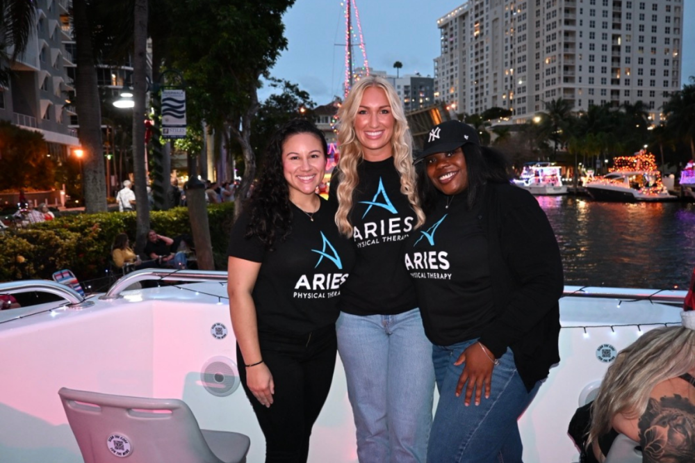 Three people smiling on a boat, wearing 'Aries Physical Therapy' shirts, with a city and decorated boats in background.