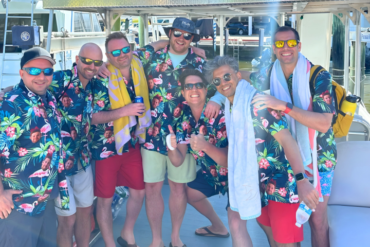 Group of seven smiling men in floral shirts posing on a boat with sunglasses and towels.