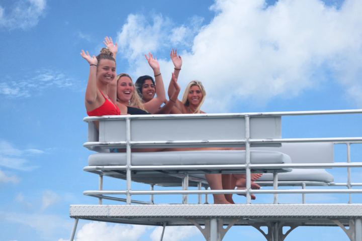 Four people waving and smiling on a boat deck against a blue sky.