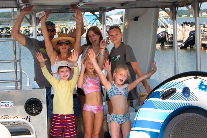 Group of smiling people on a boat by the water, some with arms raised, near inflatable tube.
