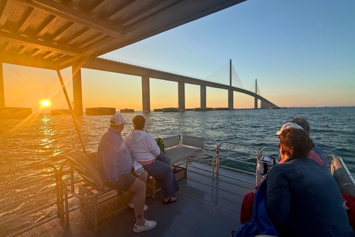 People on a boat at sunset near a large bridge over water.