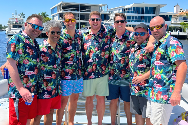 Seven men in matching tropical shirts and sunglasses standing on a boat.
