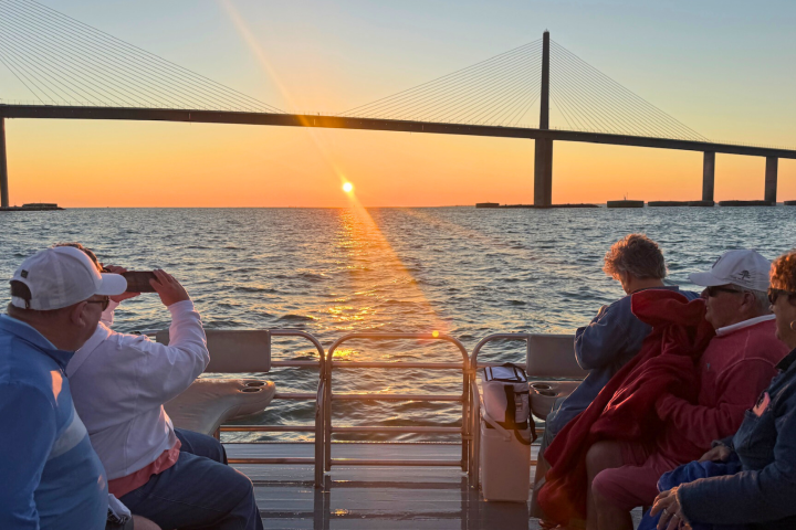 People on a boat watch the sunset near a bridge over water.