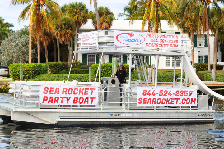 Party pontoon boat with slide, docked near palm trees and a white house.