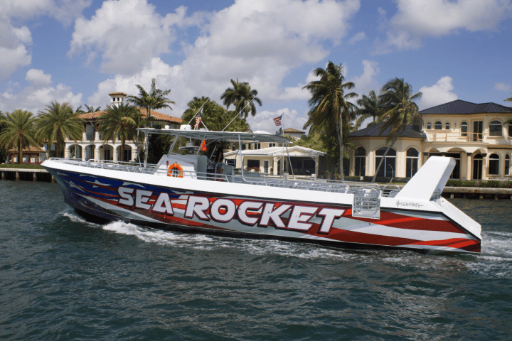 Speedboat with 'Sea Rocket' design on water, palm trees and luxury houses in background.