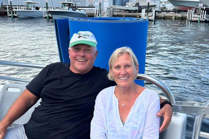 Two people smiling on a boat with water and docks in the background.