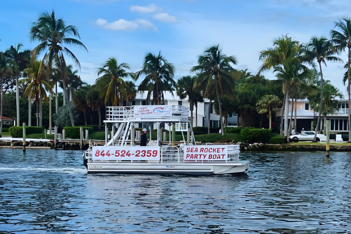 Party boat on waterway with palm trees and buildings in the background.