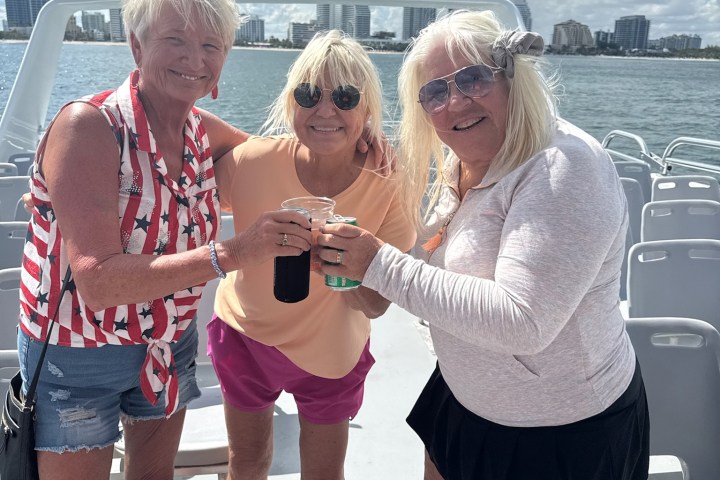 Three women smiling and toasting drinks on a boat under a cloudy sky.