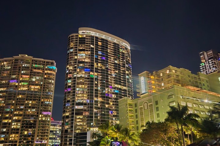 People on a boat at night in a city with lit modern buildings and palm trees.