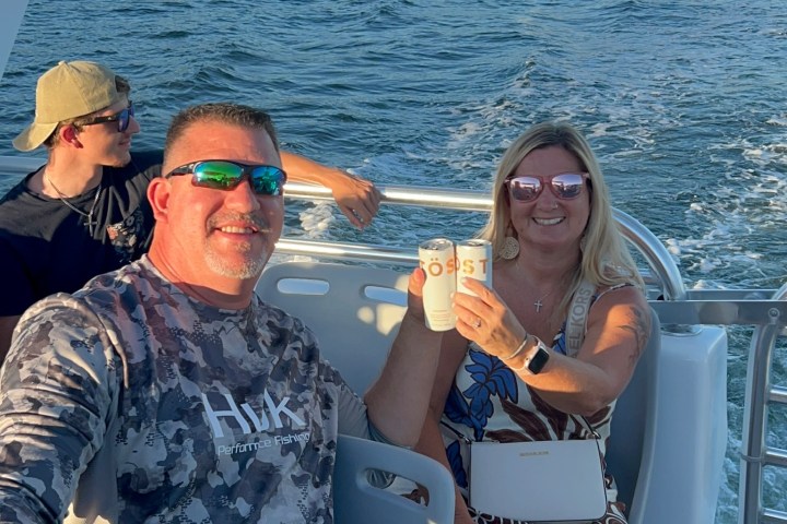 Two people on a boat holding drinks, with water and palm trees in the background.