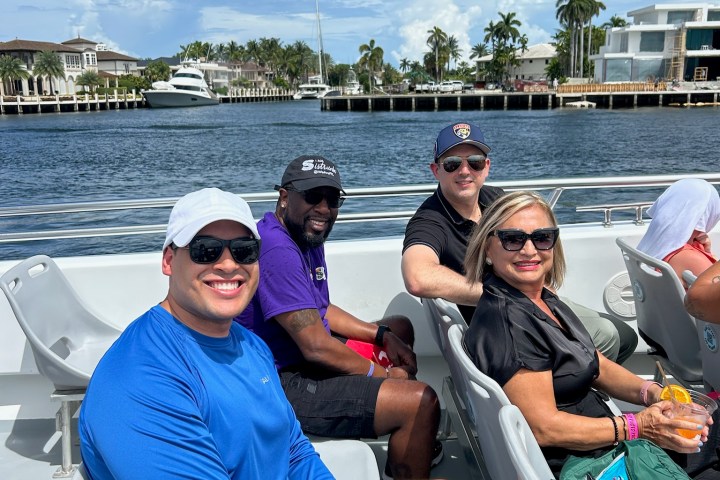 Four people enjoying a boat ride on a sunny day with water and palm trees in the background.