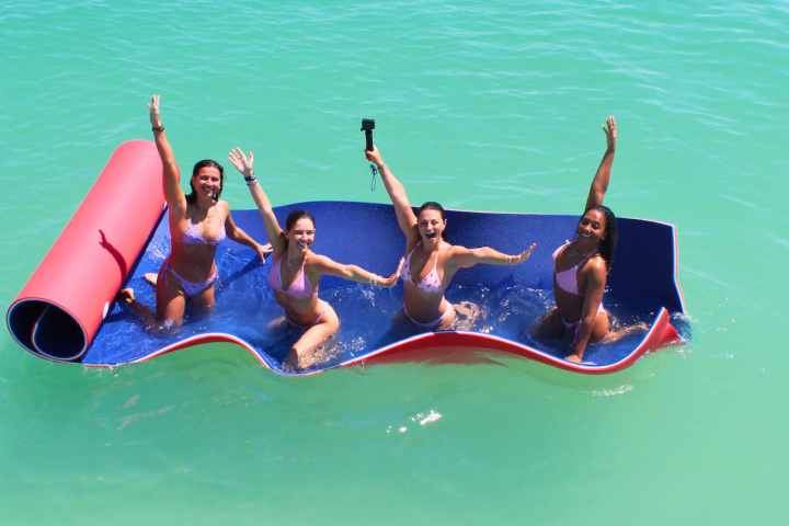 Four people on a floating mat in turquoise water, smiling and waving.