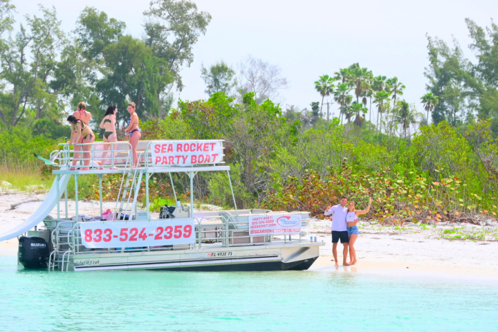 People enjoy a party boat named Sea Rocket by a sandy beach with trees in the background.