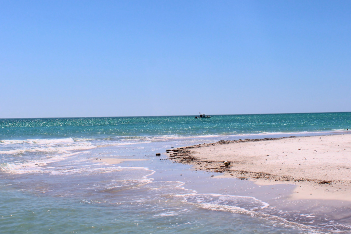Beach with gentle waves and distant small boat under clear blue sky.