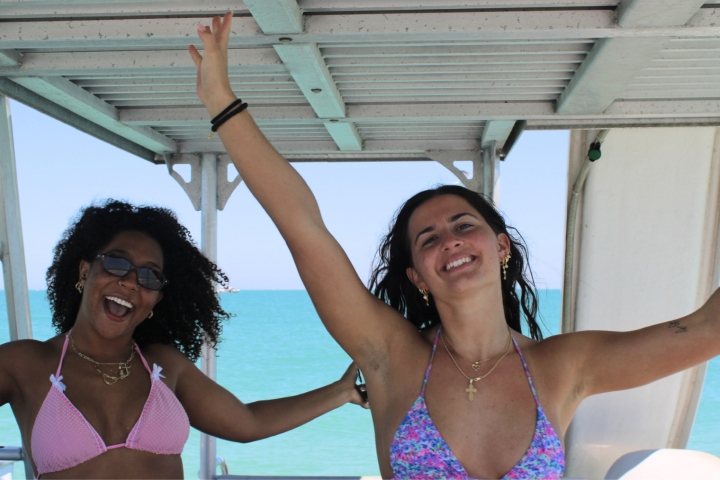 Two women in bikinis smiling and raising arms on a boat.