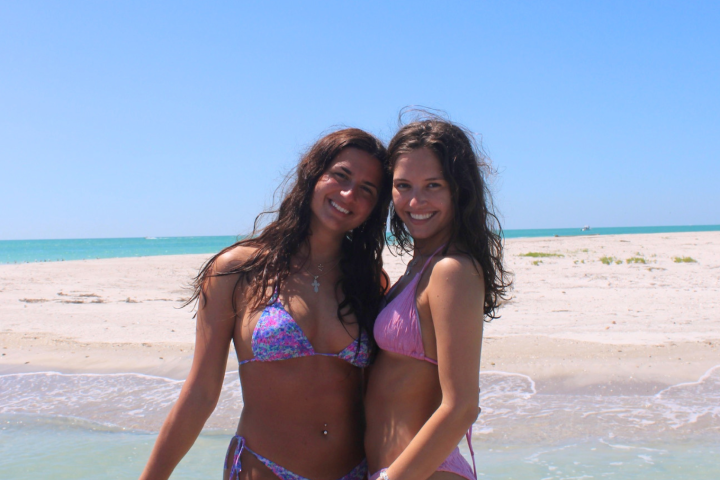 Two people in swimsuits smile on a sunny beach with ocean in the background.
