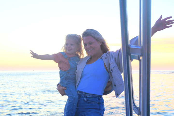 Smiling woman holding child on boat at sunset, arms outstretched.