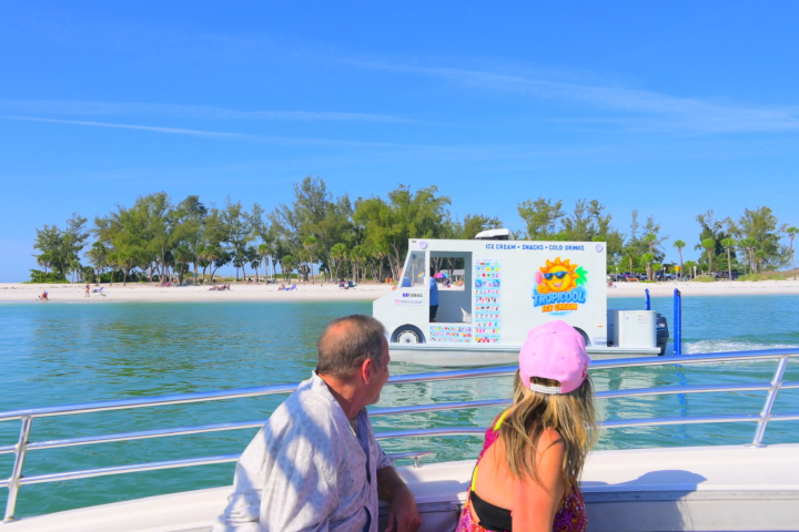 People on a boat view an ice cream truck on a sandy beach.