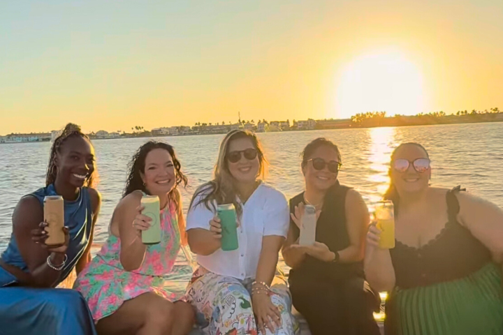 Five people sitting on a boat holding drinks with a sunset in the background.